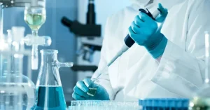 A scientist in a lab coat and gloves conducts research, using a pipette to transfer liquid into a glass vial surrounded by laboratory glassware and equipment.