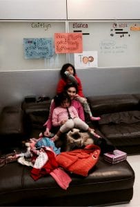 Three children sit closely together on a brown sofa, surrounded by toys, clothes, and a cat. Colorful name signs and notes posted behind them reflect a warm home focused on family wellness and togetherness.