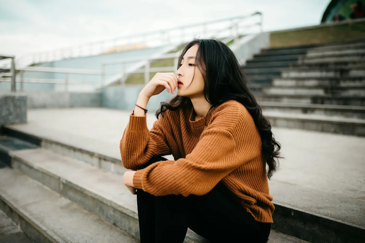 A woman with long dark hair sits on outdoor steps, wearing a brown sweater and black pants, resting her chin on her hand and looking to the side, lost in thought about educational support.