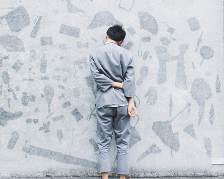 A person in gray clothing stands barefoot facing a wall covered with abstract, textured gray shapes, with one arm bent behind their back, reflecting on family wellness and the importance of emotional and educational support.