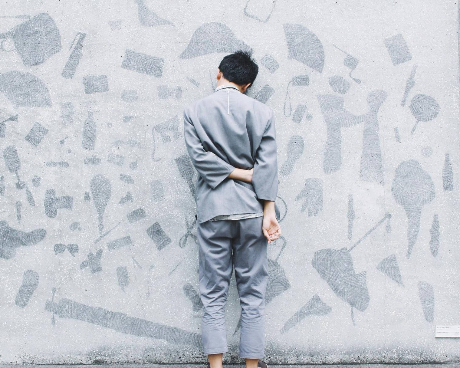 A person in gray clothing stands barefoot facing a wall covered with abstract, textured gray shapes, with one arm bent behind their back, reflecting on family wellness and the importance of emotional and educational support.