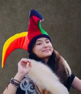 A person wearing a multicolored jester hat and fur-trimmed cloak poses and smiles in front of a plain, dark background, embodying the playful spirit and emotional wellness that curajoy encourages.