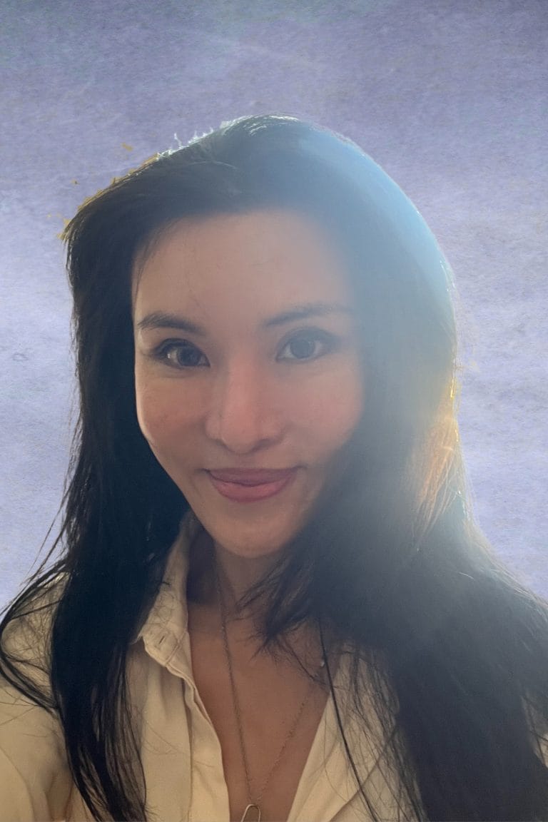 A woman with long dark hair wearing a white shirt smiles at the camera, sunlight illuminating her hair against a plain background—reflecting curaJOY’s commitment to educational support.