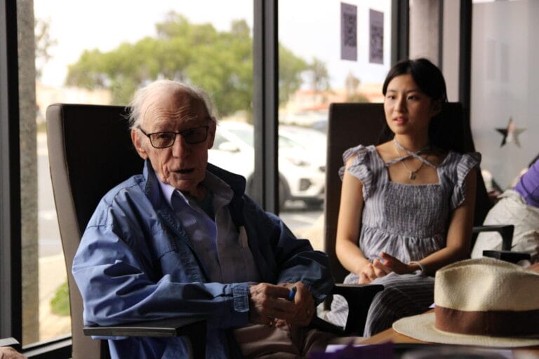 An elderly man in glasses and a blue jacket sits next to a young woman in a gray dress inside a room with large windows, reflecting a warm moment that highlights the importance of emotional wellness and connection across generations.