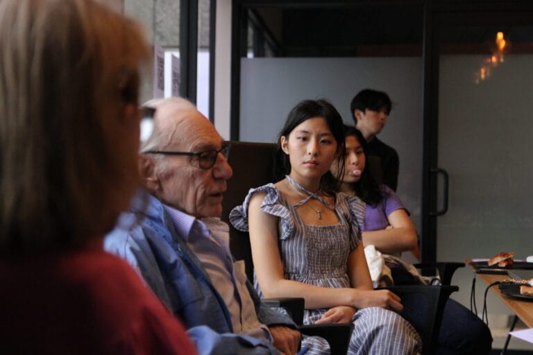 A group of people sit indoors, engaged in conversation about emotional wellness; one older man speaks while a young woman in a striped dress listens attentively, with others in the background, reflecting the spirit of curajoy.