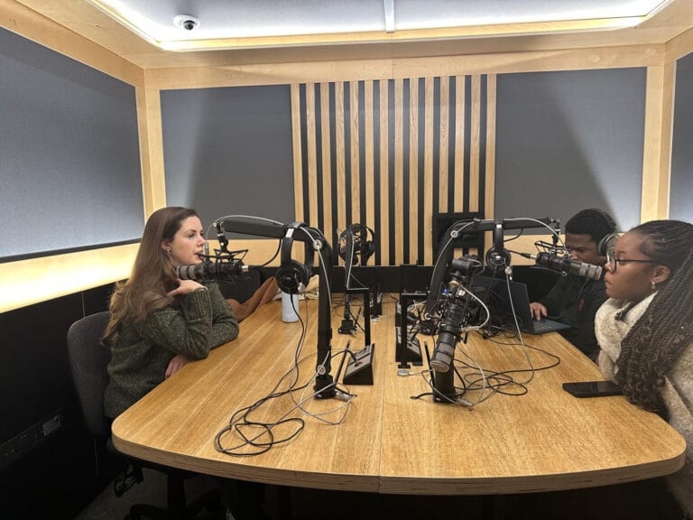 Three people sit at a table in a recording studio, each speaking into a microphone about emotional wellness. One person is typing on a laptop, while a smartphone lies on the table.