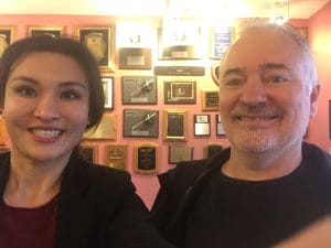 Two people smiling in front of a wall covered with several award plaques, celebrating their innovative product design philosophy.
