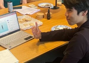 A person sits at a table with cookies, a laptop showing a 3D character, and paperwork, making a hand gesture with two fingers raised—capturing a moment that highlights emotional wellness.