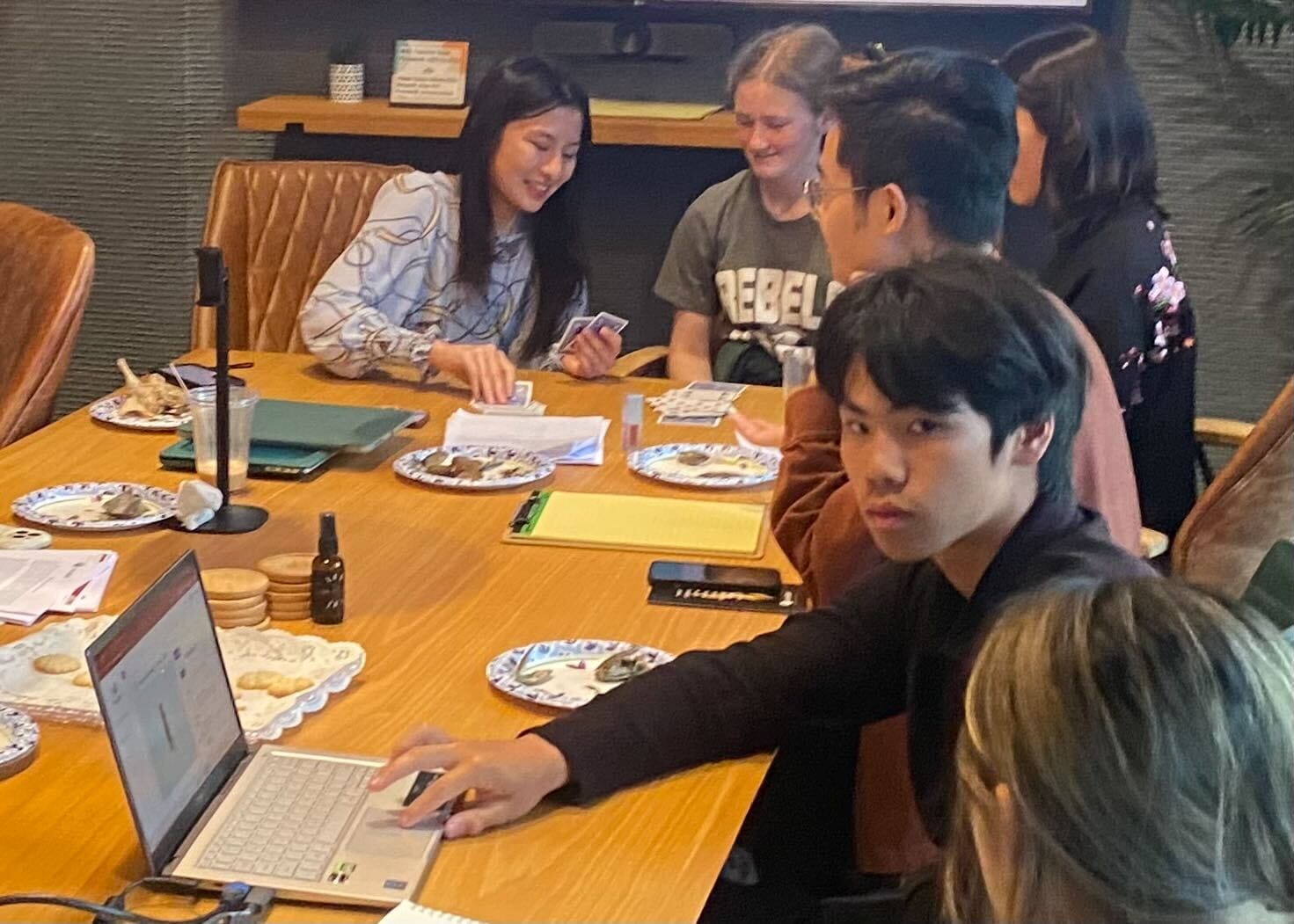 A group of people sit around a table with laptops, papers, and snacks, some discussing youth mental health and others working, in a casual meeting room setting focused on curaJOY and educational support.