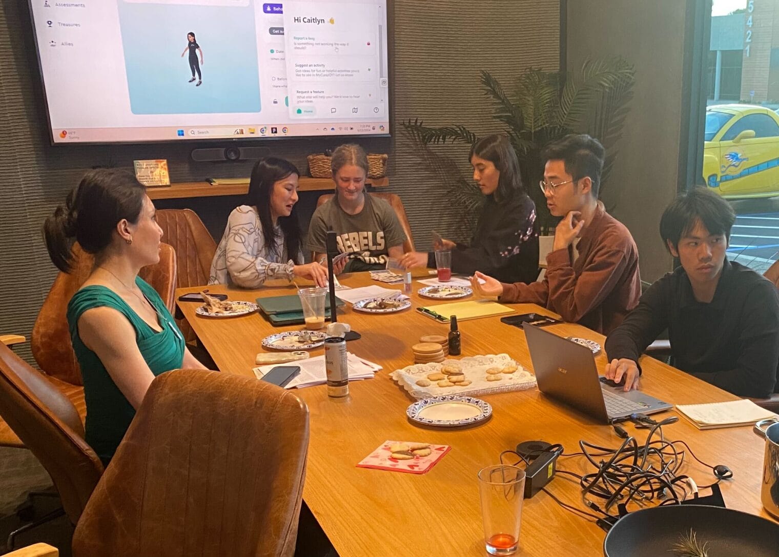 Six people, participating in an internship, sit around a conference table with laptops, snacks, and drinks, while a screen displays a presentation in the background.