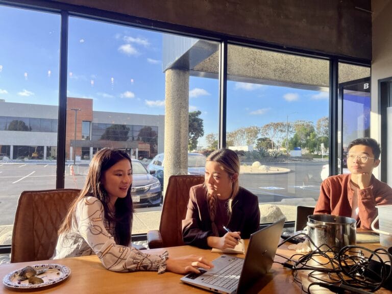 Three people sit at a table in a sunlit office, engaged in discussion about curajoy and emotional wellness with a laptop open; a parking lot is visible through large windows behind them.
