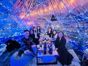 A group of people sits around a table with food and drinks inside a transparent dome decorated with blue lights, enjoying a warm atmosphere that encourages family wellness.