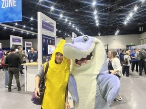 Two people in costume, one as a banana and the other as a shark, pose together at a busy convention promoting curaJOY and responsible technology for youth mental health.