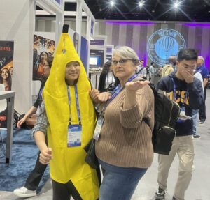 Two people pose at a convention; one is wearing a banana costume while the other stands beside them. Attendees walk by in the background, passing booths and displays about curajoy and behavioral healthcare.