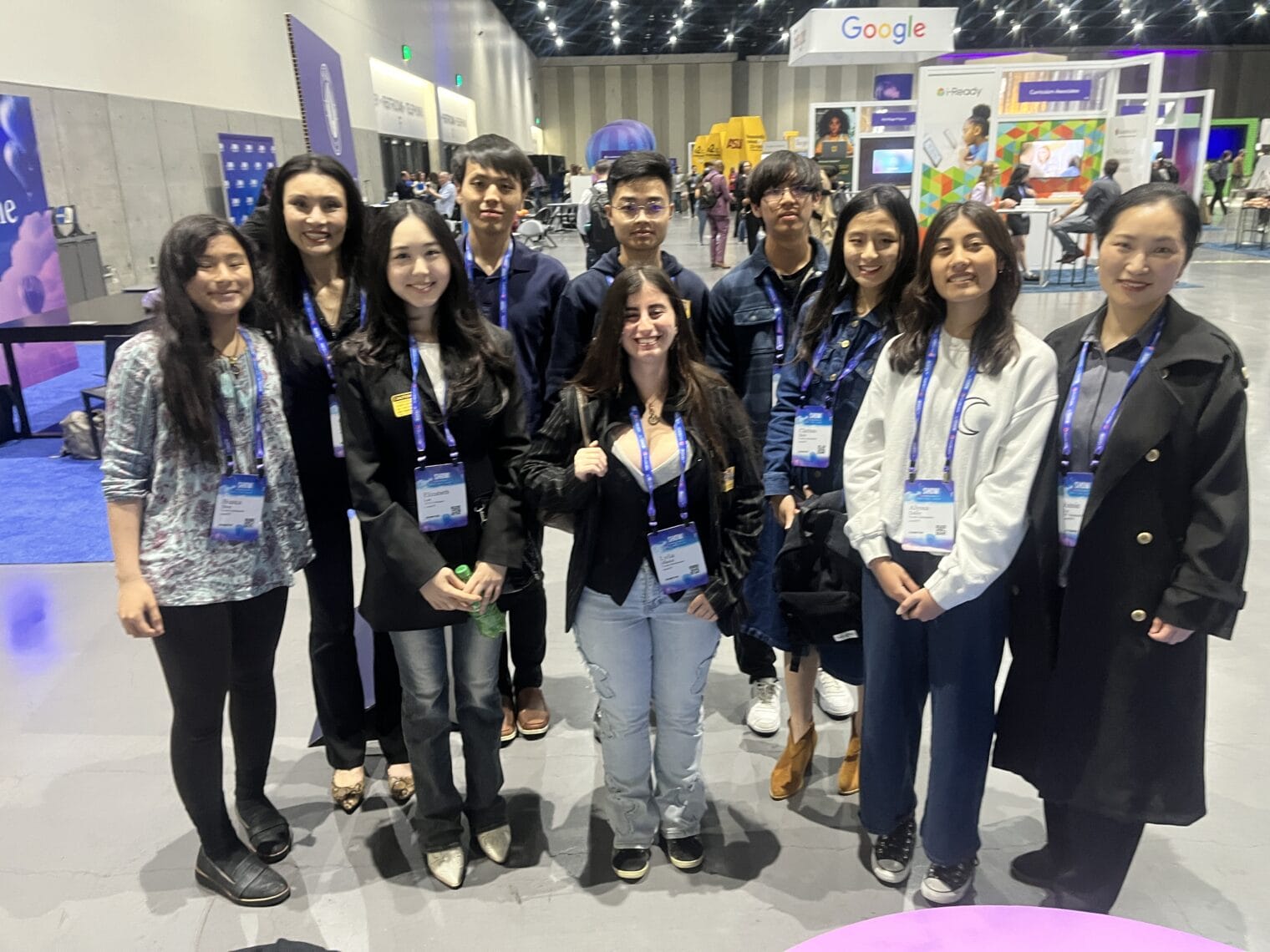 A group of ten people wearing conference badges pose together indoors at a tech event, with booths and displays visible in the background.