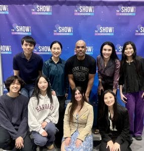A group of nine people pose in front of a blue backdrop with "THE SHOW" logos. Smiling and casually dressed, they gather to promote youth mental health and provide educational support, reflecting the curaJOY mission.