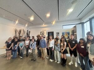 A group of people, mostly young adults and teenagers, pose for a photo in an art gallery with various framed artworks—some labeled "Ready to Ship"—displayed on the walls behind them.