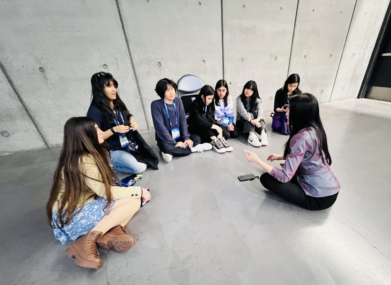 A group of seven people sits in a circle on a concrete floor near a gray wall, listening as a youth ambassador speaks to them, fostering discussion and youth engagement.