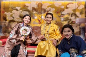 Three people pose indoors in traditional East Asian clothing, with an ornate gold decorative mural in the background, highlighting curaJOY's educational support for cultural appreciation.