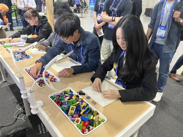 Two people sit at a table sorting and assembling colorful LEGO bricks on base plates, with containers of LEGO pieces in front of them; others observe in the background, creating a vibrant space for youth engagement.