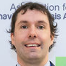 A person with wavy brown hair and a light complexion is smiling at the camera in front of a blurred sign with partially visible text.