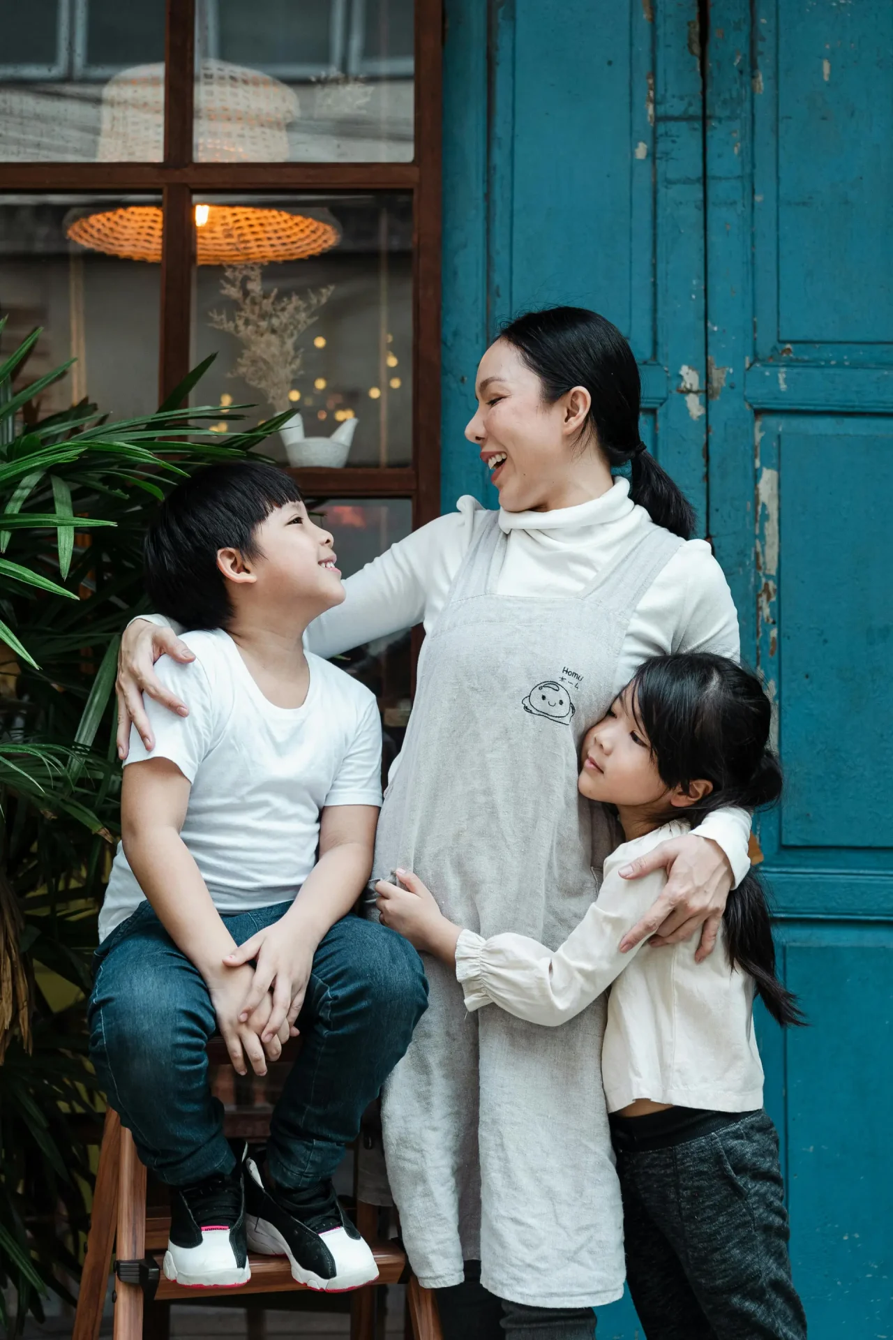 A woman stands smiling with her arms around a young boy and girl, all looking at each other, in front of a blue door and plants.