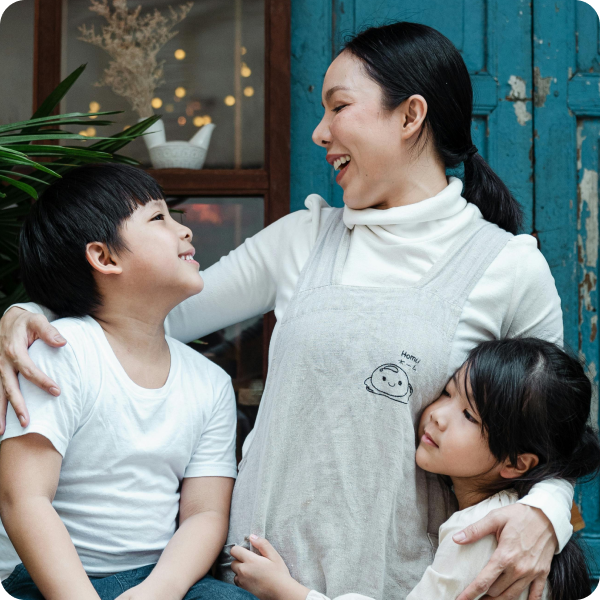 A woman sits smiling with two children, one sitting beside her and the other hugging her, in front of a blue wooden door.