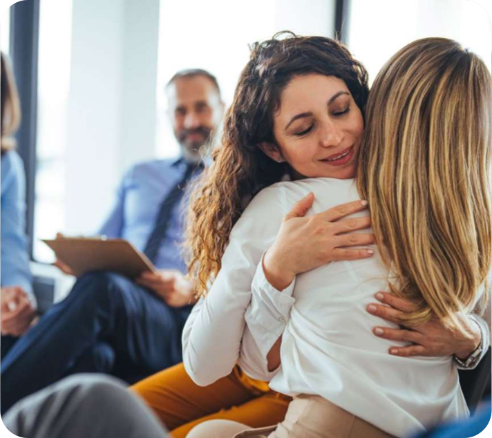 Two women are hugging each other in an indoor setting, while a man with a clipboard sits in the background, suggesting a support or therapy group environment.