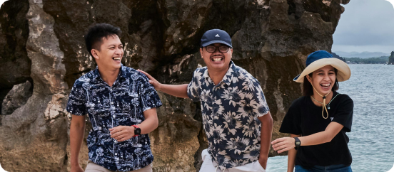 Three people wearing patterned shirts and hats smile and laugh together while standing by rocky cliffs near the ocean.