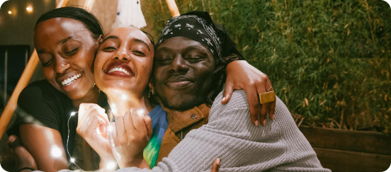 Three people sit close together, smiling and hugging, with one person holding string lights; they appear happy and relaxed in an indoor setting with greenery in the background.