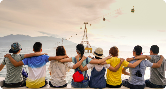 Seven people sit side by side on a ledge by the water, arms around each other, facing away, with cable cars visible in the background over the sea.