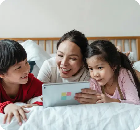 An adult and two children lie on a bed, smiling and looking at a tablet together, enjoying responsible technology that supports youth mental health.