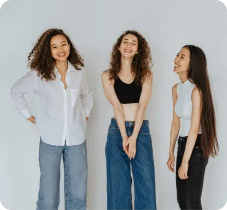 Three women stand together smiling and laughing against a plain light-colored background, wearing casual tops and jeans, showing the positive impact of strong behavioral health and supportive friendships.
