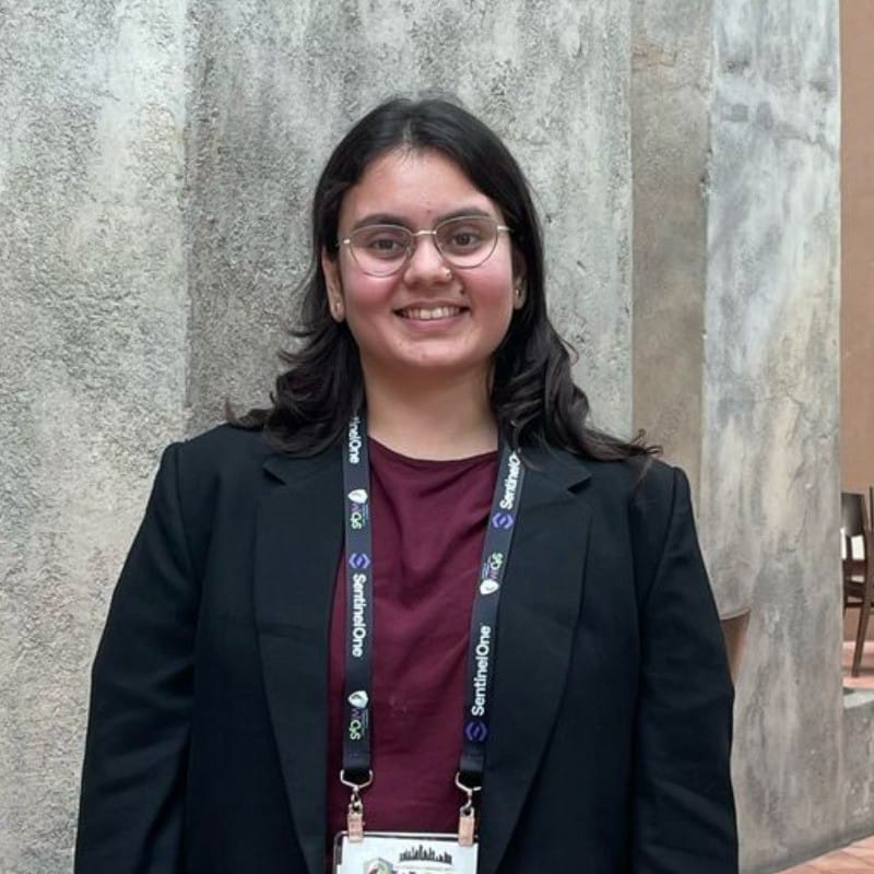 Safe to Break, Ready to Ship | curaJOY A person wearing glasses, a black blazer, and a "Ready to Ship" conference lanyard stands and smiles in front of a textured wall.