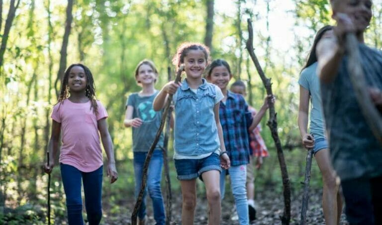 Diverse group of children walking on a forest trail, with one girl in the center giving a thumbs up.