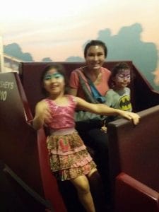 A woman sits with two children in face paint on a theme park ride, enjoying family wellness; one child wears a pink outfit and the other sports a Batman shirt.