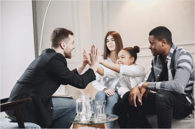 Four people sit on a couch, smiling and engaging in a high-five. There are two glasses and a water pitcher on the table in front of them.