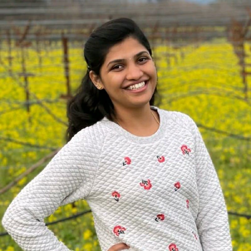 Safe to Break, Ready to Ship | curaJOY A woman with long dark hair, wearing a light-colored sweater with small floral prints, stands smiling in front of a field with yellow flowers and rows of bare vines—her look is Ready to Ship for any spring day.