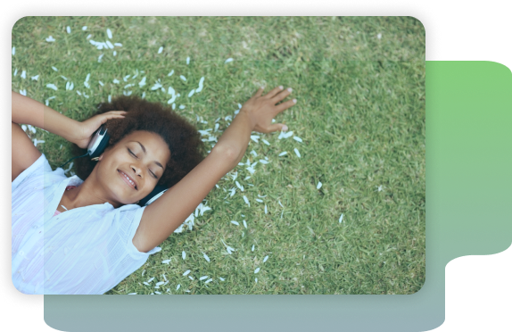 A person lies on grass with petals around them, eyes closed and smiling, wearing headphones and a white shirt.