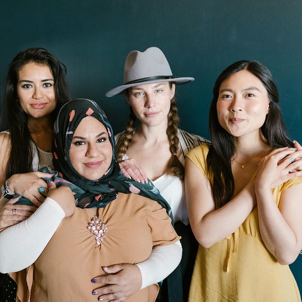 Four women stand together in front of a dark blue wall, smiling at the camera. Representing curaJOY’s commitment to family wellness, they showcase diversity in appearance and dress, with one wearing a headscarf and another a hat.